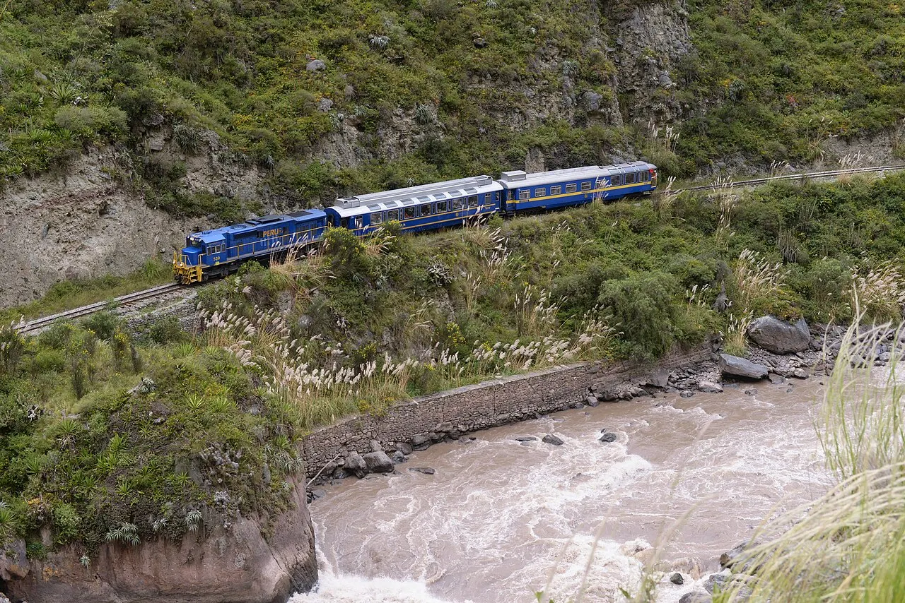 A blue PeruRail train threading the gorge above the churning Urubamba river toward Machu Picchu